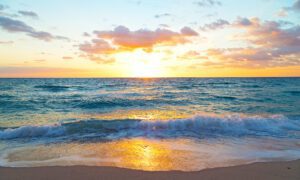 The beach at sunset, where the sky is several shades of orange and yellow and the shore is sandy brown with foamy blue waters touching it.